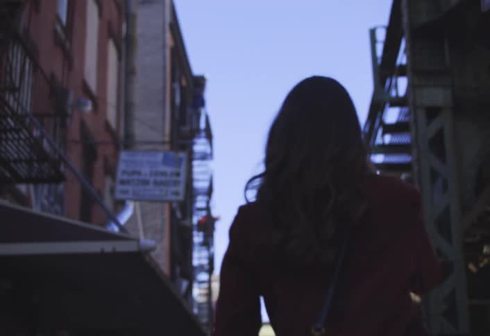 Closeup back shot of a woman walking through an urban street with city buildings and a metal staircase in the background