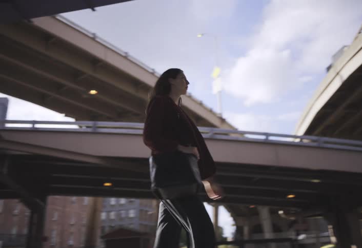 Walking on a city street beneath multiple elevated highway overpasses a woman wearing a jacket and carrying a bag looks upward with background buildings and vehicles visible