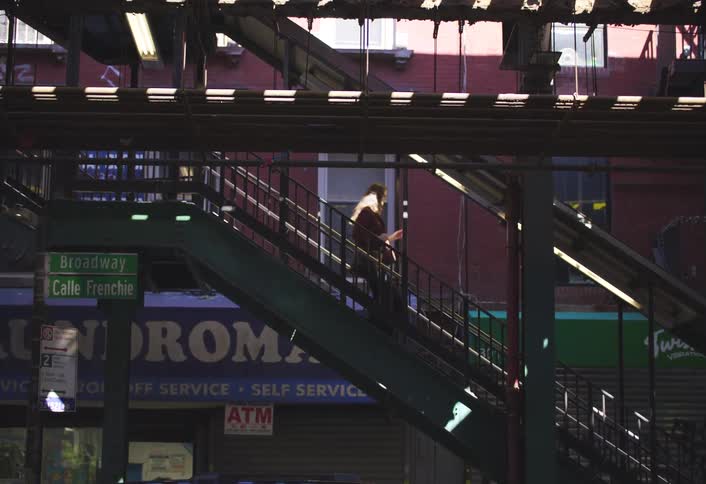 A woman walks down the stairs of a city subway station near a broadway street sign