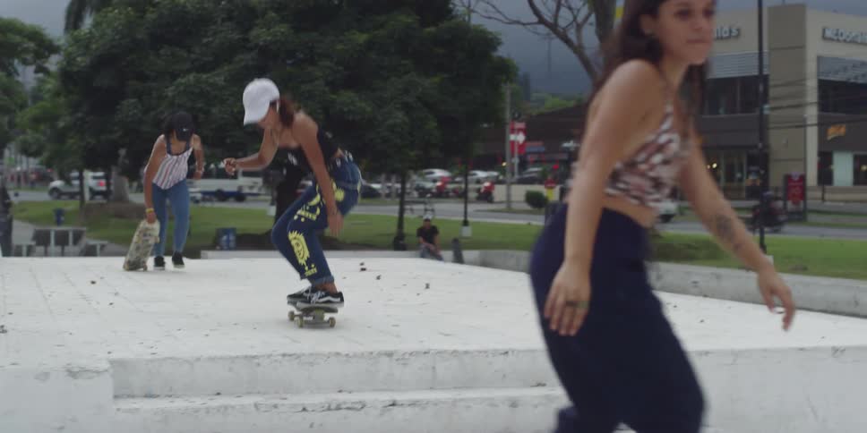 Female skateboarders jump their skateboards down the outdoor steps of a building near the street in a commercial area under a gray sky
