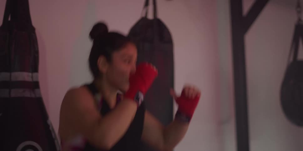 A person in red gloves executes a high kick in a gym surrounded by punching bags