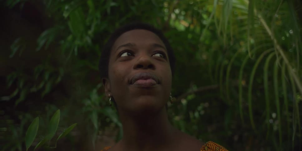 A young black woman with short hair looks up and smiles against a backdrop of lush green foliage
