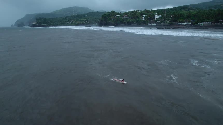 Woman paddling on a surfboard in rough seas with a strip of beach and hills covered in vegetation in the background