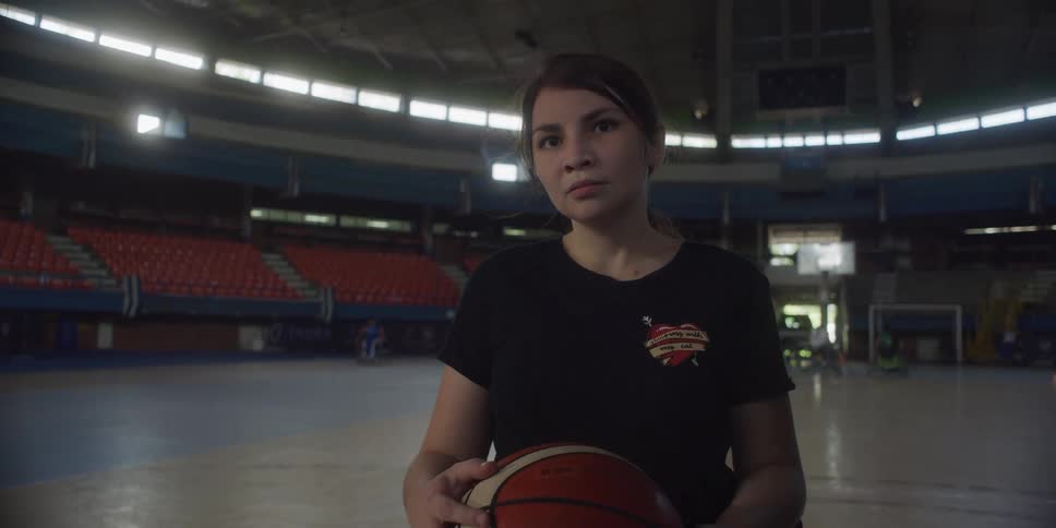 A person in a wheelchair holds a basketball on an indoor court with red stadium seating and floor cones arranged nearby