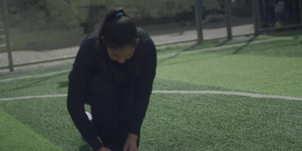 A woman wearing black exercise clothes ties her shoes and stands up on a turf field