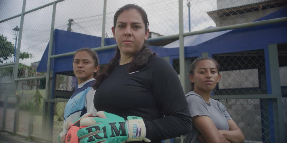 Three women are standing inside a fence of a baseball field and one has gloves on holding a soccer ball