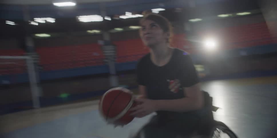 Two disabled women in wheelchairs are playing basketball on a court one passes the ball while the other shoots it into the basketball hoop