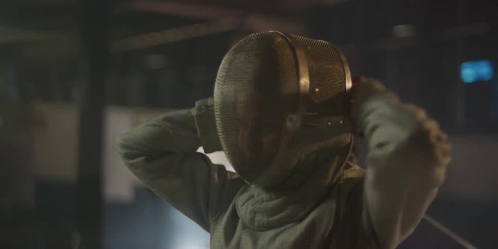 A teenage girl inside a court wearing a fencing mask and holding a fencing sword poised and ready to begin fencing