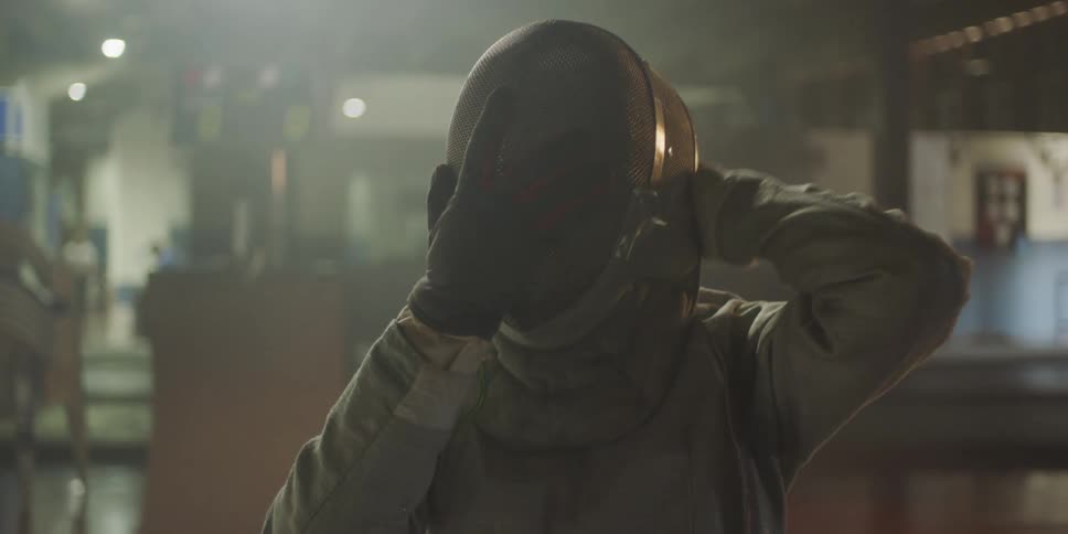 A young woman puts on a fencing helmet while sitting indoors