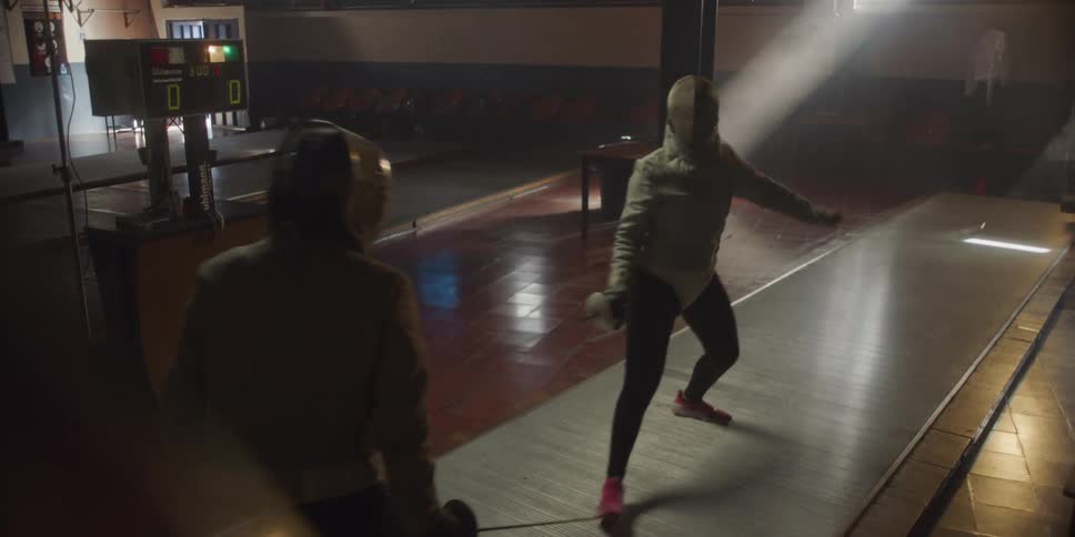 Two women are fighting in a fencing match in a dimly lit room with a scoreboard