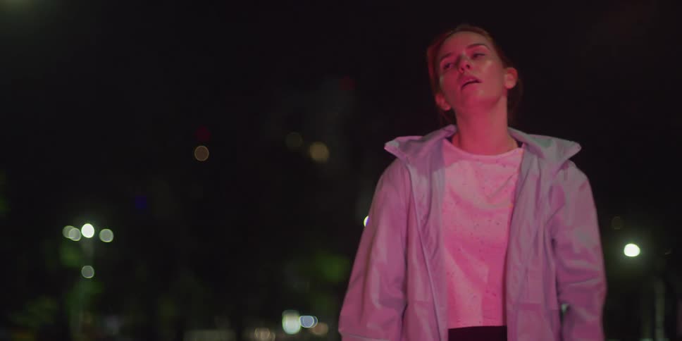 A young woman is running alone at night on the street and comes to a stop looking tired she is wearing a jacket and a white tshirt