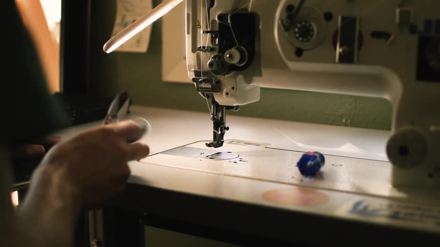 A person with visible forearm tattoos is carefully sewing a dark piece of fabric using a well lit industrial sewing machine
