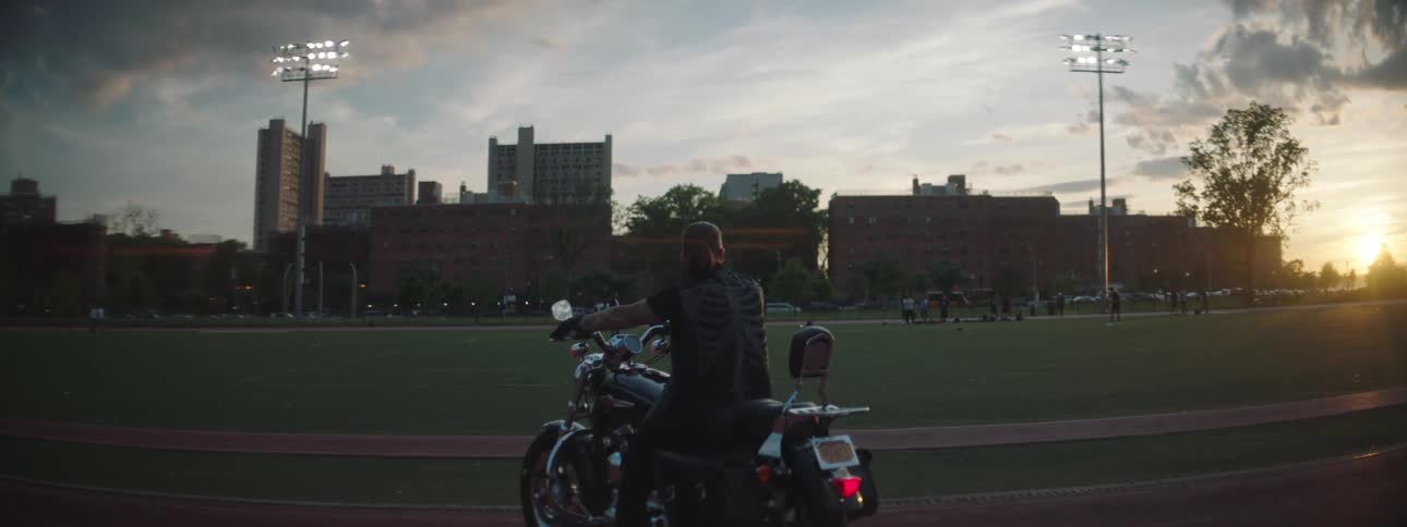 Person sitting on a motorcycle parked next to a floodlit sports field with residential buildings in the background and the sun setting on the horizon