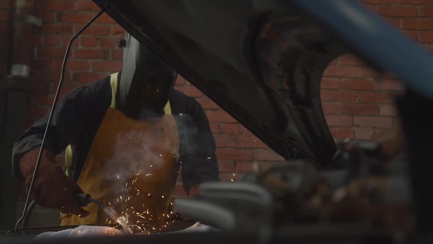 Welder working inside the hood of a car with sparks visible wearing protective gear