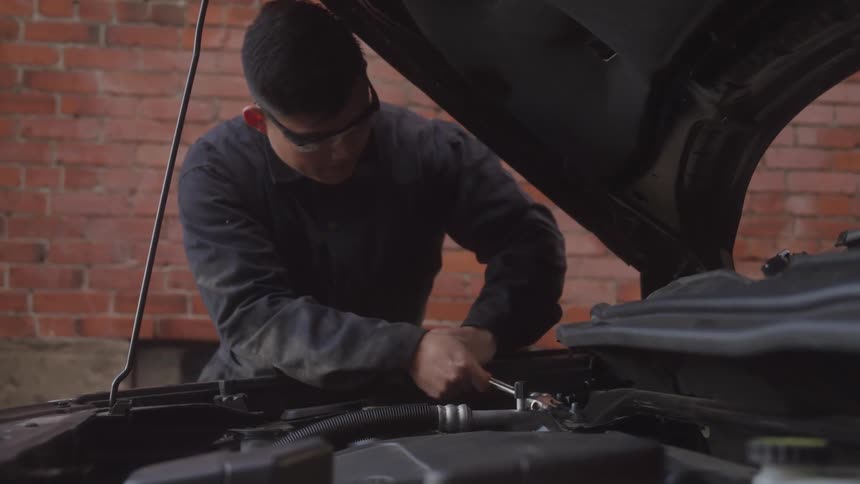 Mechanic working on a car engine with the hood open in front of a brick wall