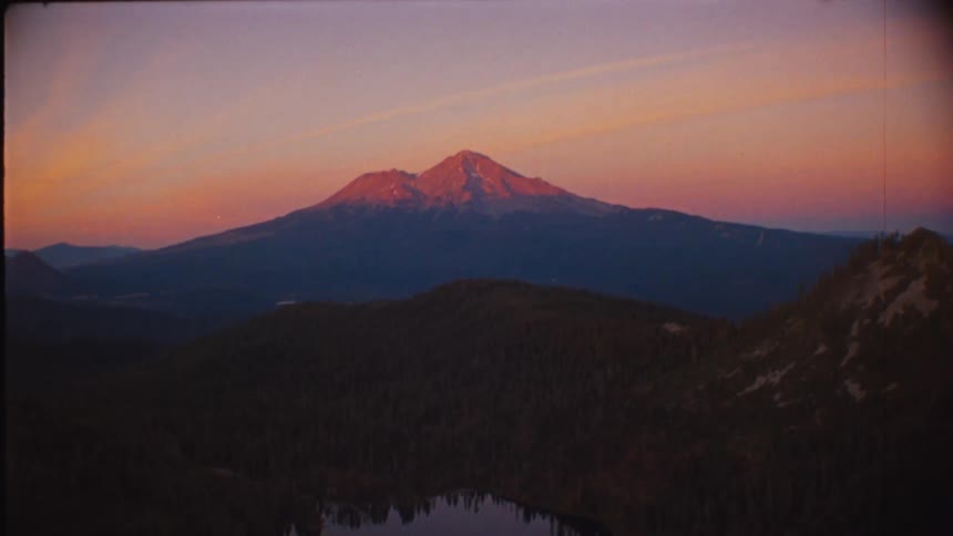 Shirtless man stands near the edge of a cliff and looks at the mountainous terrain surrounding a lake under a red sky