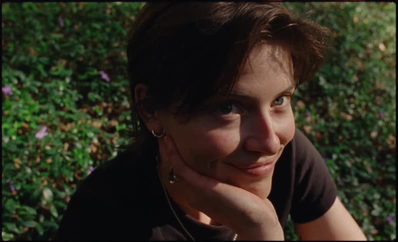 Woman with a smiling expression resting her face on her hand in the middle of an outdoor environment with green vegetation and small purple flowers in the background