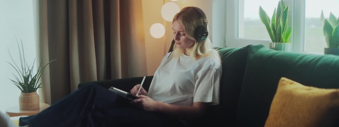 A woman with headphones sits on a green couch writing in a notebook with a plant on the side table and three round lights in the background