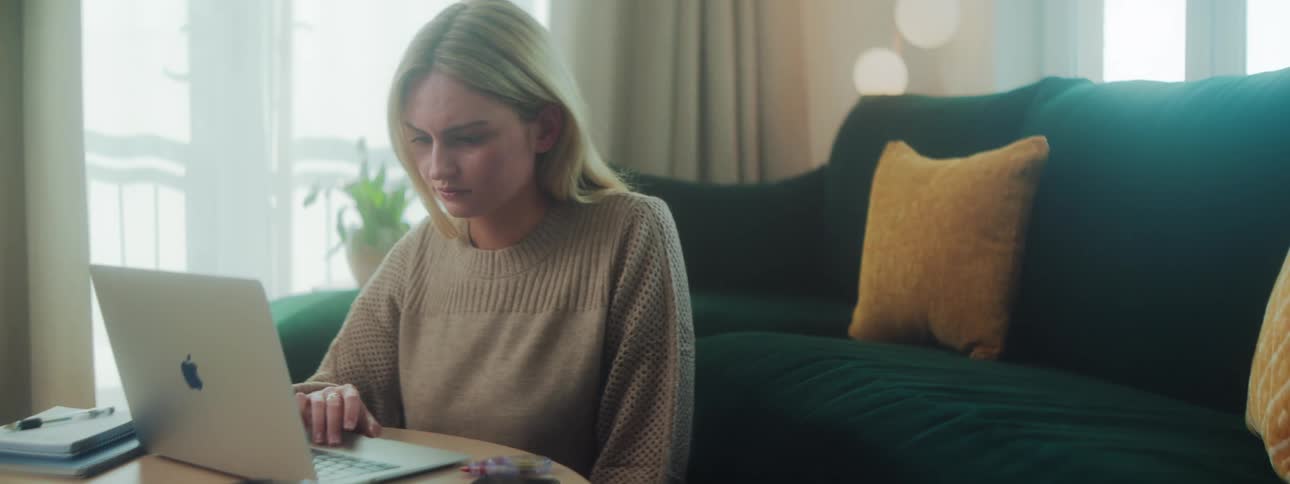 The face of a young blonde woman who is sitting next to the sofa and using a laptop