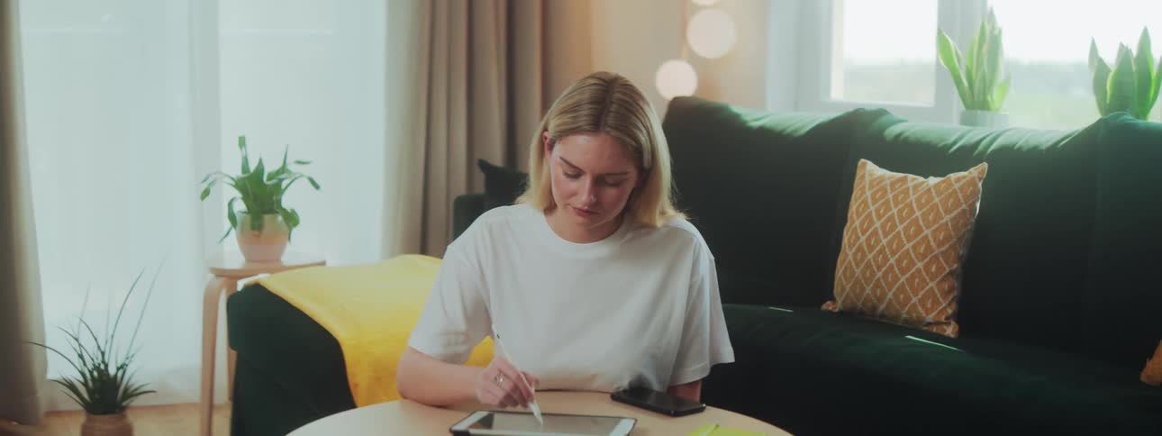 A woman sits at a round table in a cozy living room using a stylus on a tablet surrounded by cushions plants and a smartphone