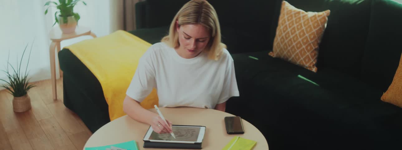 A woman in a room sitting in front of a table drawing on her tablet