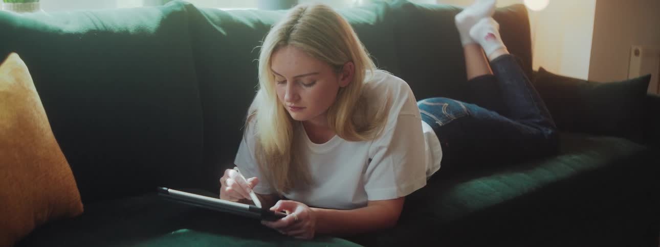 A woman is drawing on a tablet while laying on the couch
