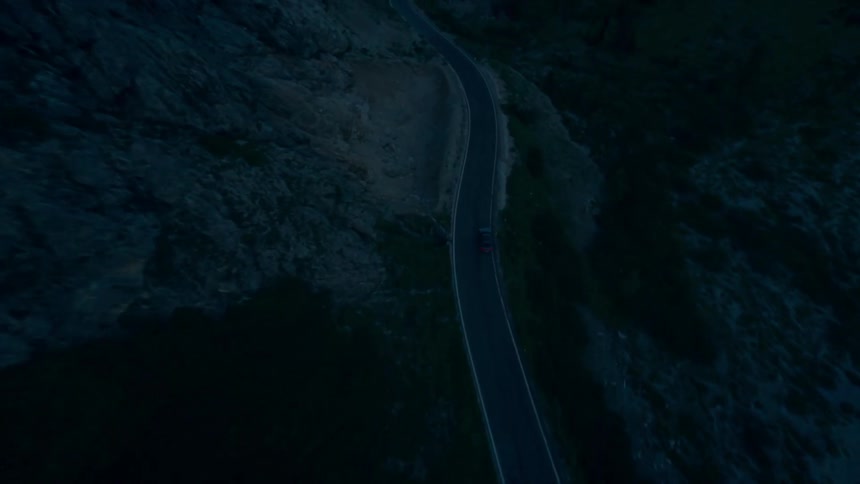 A black sedan drives along a coastal road surrounded by mountains with the warm glow of sunset casting light over the scene