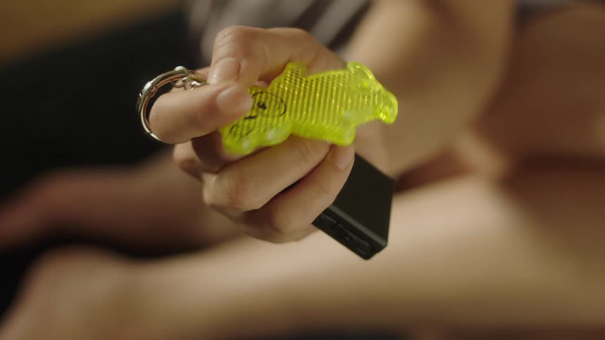 A close up of a hand holding a yellow glossy cartoon bear shaped keychain attached to a black box possibly a controller via a silver keyring and chain