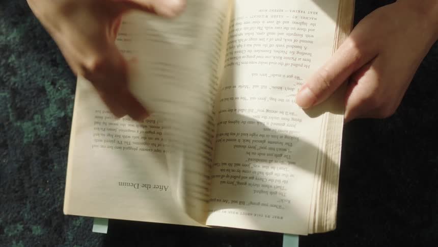 Two hands flip through the pages of an open book bathed in warm sunlight with blue page markers visible