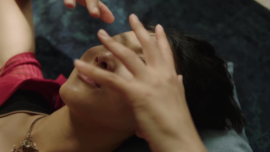 A person lies on a dark blue rug with arms stretched above their head wearing a red ribbed shirt a gold necklace and resting on a gray pillow