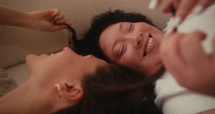 Two women lying on a couch laughing as they playfully touch and run their fingers through each others hair