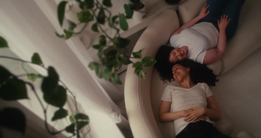Two people relax on a beige couch amidst lush green plants dressed in lightcolored clothing