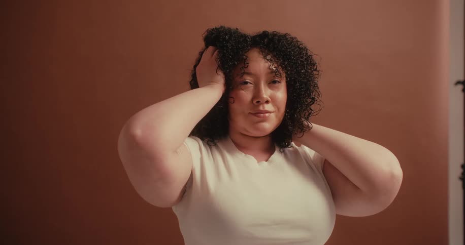 I woman touches her curly brown hair while standing in front of a brown backdrop