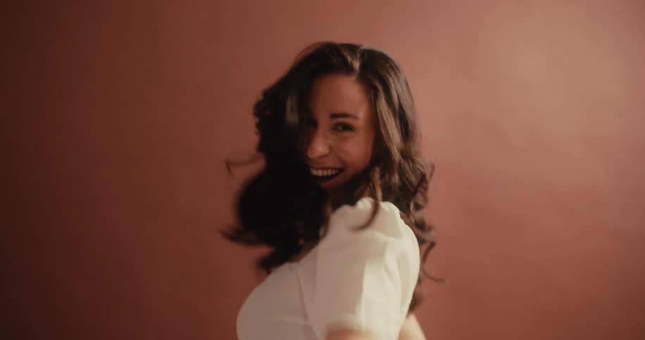 Closeup of a smiling woman with wavy long brown hair modeling a white blouse against a brown backdrop while looking at the camera