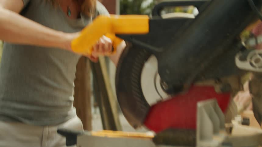 A woman is using a power saw to cut wood in front of a house