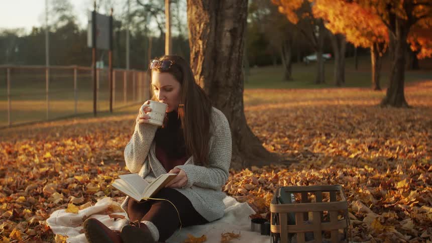 A young woman sitting in a park in autumn holding a book in one hand and drinking coffee with the other