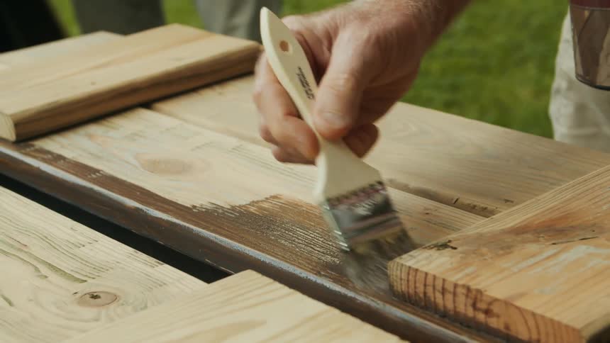 A person applies dark wood stain to wooden planks using a paintbrush