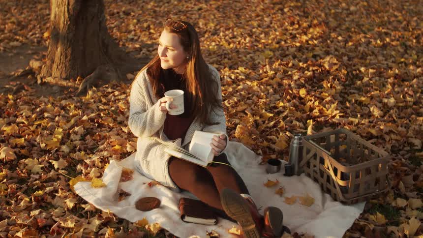 A woman sits on a blanket in a park surrounded by vibrant autumn leaves holding a cup with a book a basket and a thermos nearby