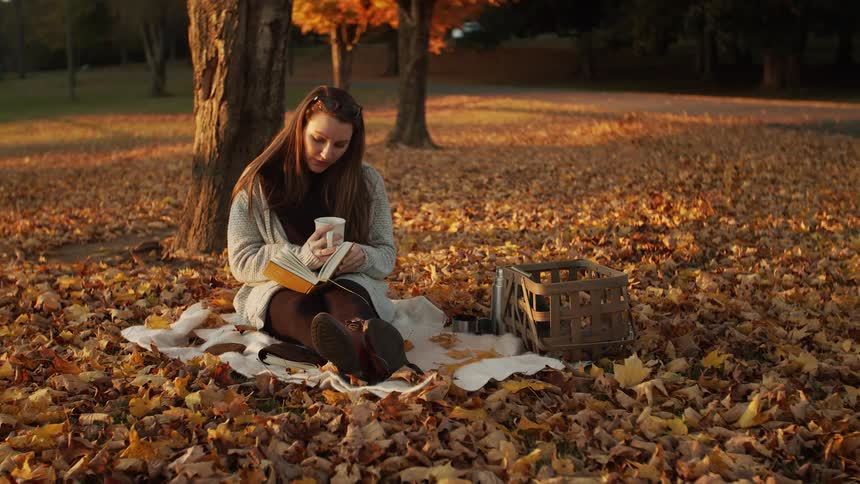 A woman is drinking from a mug while reading a book in the park