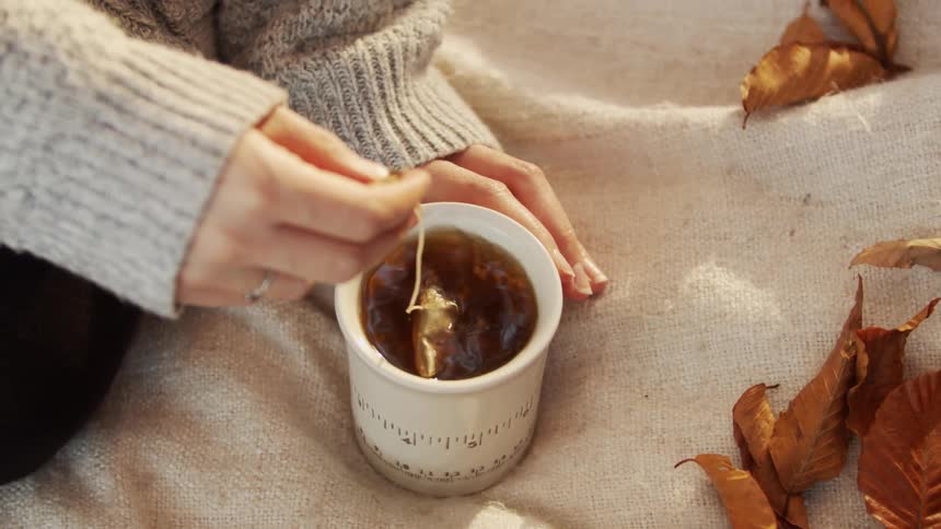 A tea bag is being dipped on a cup of tea on a beige fabric blanket surrounded by dried autumn leaves