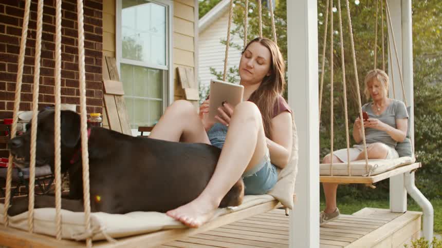 A mother and daughter sit on porch swings while looking at their smartphone and tablet
