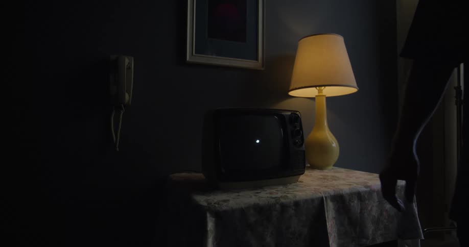 In a dimly lit room a hand turns the dial to switch off an old crt television displaying static leaving the screen dark