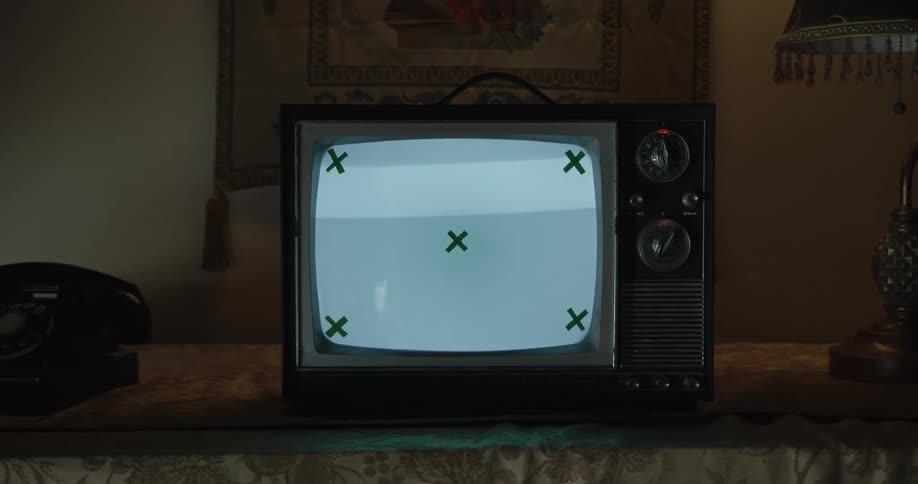 An old television displaying a blank screen with green x marks in the corners and center sits on a table next to a rotary dial telephone