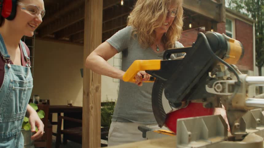 Mother and daughter wearing safety glasses and headphones operate a saw on the porch of their home