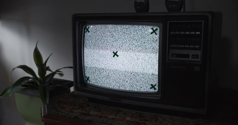 An old television with static and green x marks on its screen rests on a patterned tablecloth beside a small plant