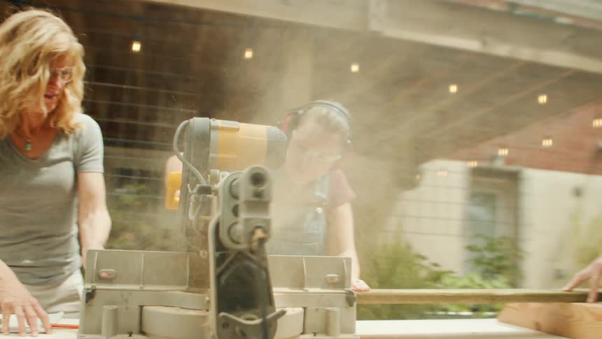 A teenager using a miter saw to cut a wooden plank in the house yard with family nearby