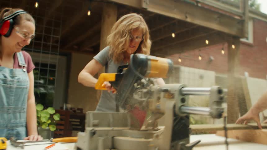 A woman is cutting wood with a power saw outside a house while others watch