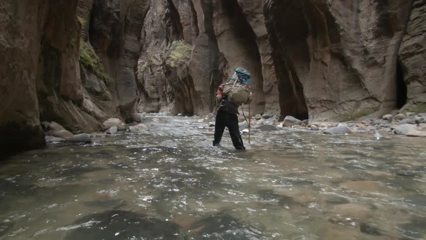 A hiker carrying a heavy backpack wades through a stream in a canyon