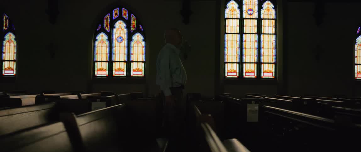 A person walks down the aisle between pews in a dimly lit church adorned with vibrant stained glass windows