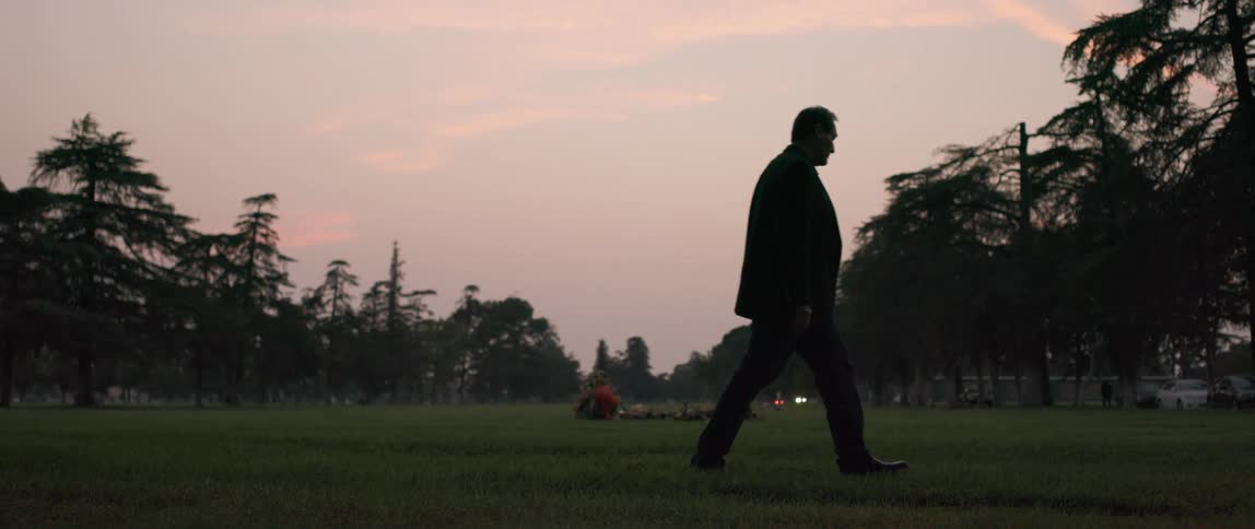 A man in a suit is walking by a grave in a cemetery at sunset